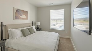 Une chambre à coucher comprenant un lit, une table de chevet, une lampe, une fenêtre avec des stores et une œuvre d’art accrochée au mur.