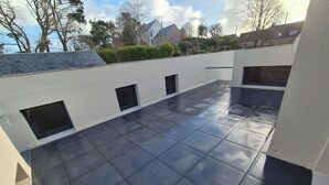 Una terraza de techo plano con baldosas oscuras, ventanas pequeñas y vistas de edificios y árboles circundantes.
