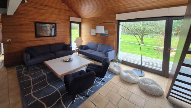 Una sala de estar acogedora, con techo de madera, dos sofás, una mesa de centro y una lámpara. Amplias ventanas ofrecen vistas a un jardín.