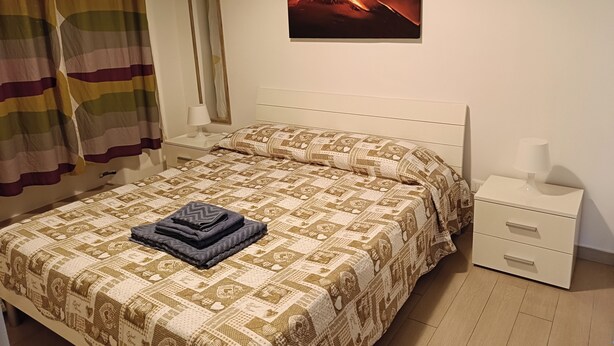 Un lit bien fait, avec une couvre-lit à motifs, une couverture pliée et un oreiller. Il y a une table de chevet avec une lampe et un tiroir. Un tableau encadré représentant un volcan est accroché au mur.