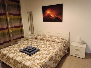 A neatly made bed with a patterned bedspread, a folded blanket, and a pillow. There is a nightstand with a lamp and a drawer. A framed picture of a volcano is on the wall. - Beautiful loft (for 3 people) in Catania, available for short-term rentals (Catania)