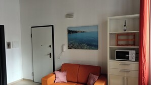Une pièce avec une porte blanche, un tableau encadré représentant un paysage côtier, un meuble blanc contenant un four à micro-ondes, et un canapé orange avec deux coussins.