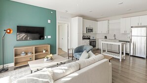 Een moderne woonkamer met een bank, salontafel en televisie. Grenzend daar aan bevindt zich een keuken met witte kasten, een magnetron en een koelkast.