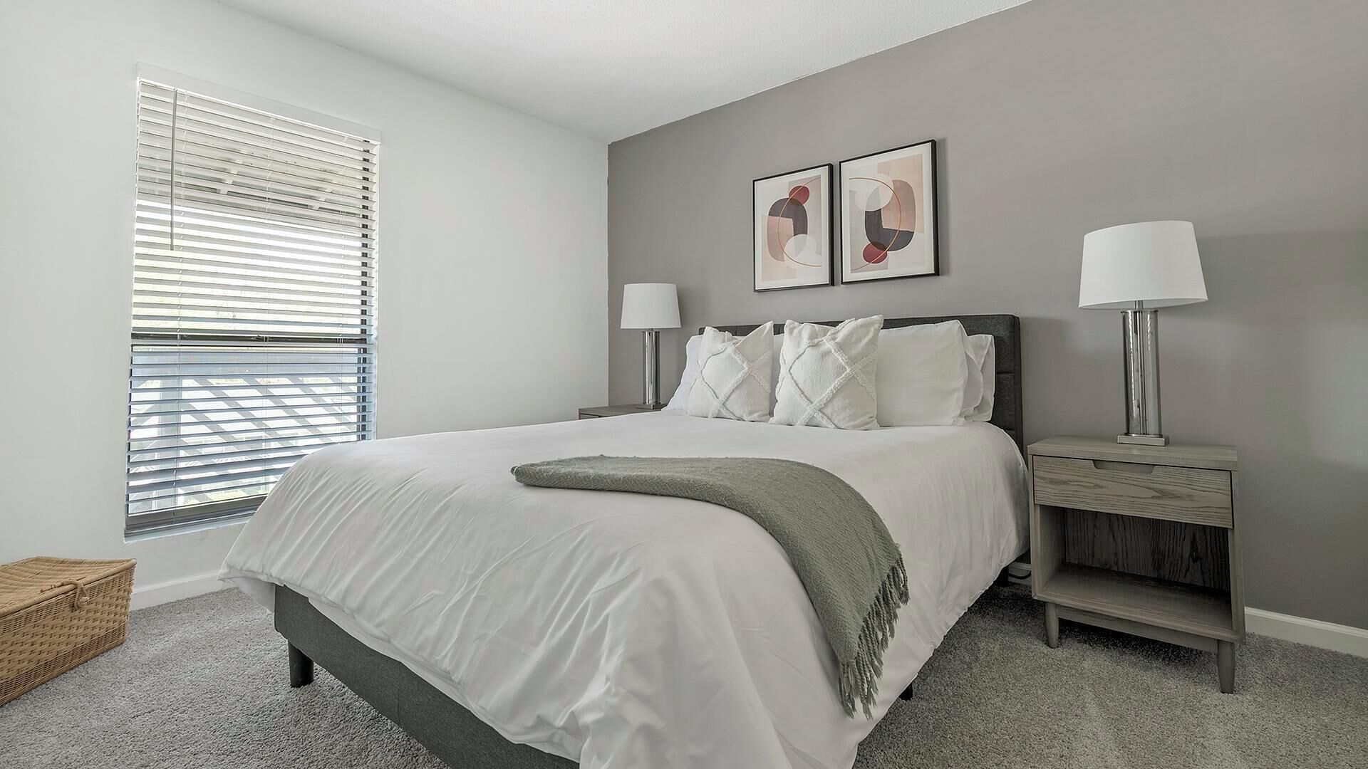 A bedroom with a bed, two bedside tables with lamps, a window with blinds, and two framed pictures on the wall.