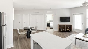 A modern kitchen with white cabinetry, a dining area with a white table and chairs, a flat-screen TV mounted on the wall, and a wooden floor. - Landing | Chic 1BD, Courtyard, Yoga Studio (Huntsville)