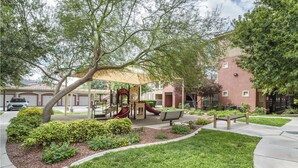 A playground with slides and a bench, surrounded by well-maintained grass and shrubs, with a building in the background. - Landing | Stunning 1BD, Gym (North Las Vegas)