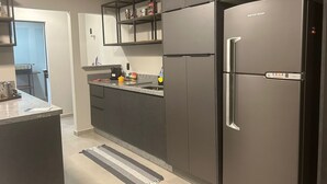 A modern kitchen with dark cabinetry, a stainless steel refrigerator, and a countertop with various items. - GEMINADO CASA 2 ALTO PADRÃO 3 QUARTOS (Joinville)