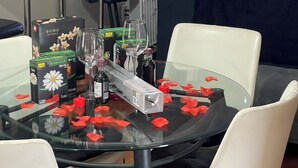 Una mesa de comedor con cubierta de vidrio, rodeada de sillas blancas, decorada con pétalos de flores rojas, copas de vino y una botella.