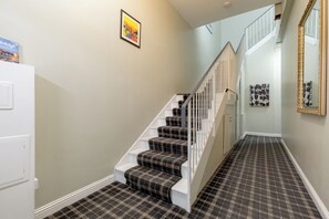 Un escalier recouvert d’un tapis à carreaux et doté de rampes blanches, menant à une pièce avec une porte blanche et un tableau encadré accroché au mur.