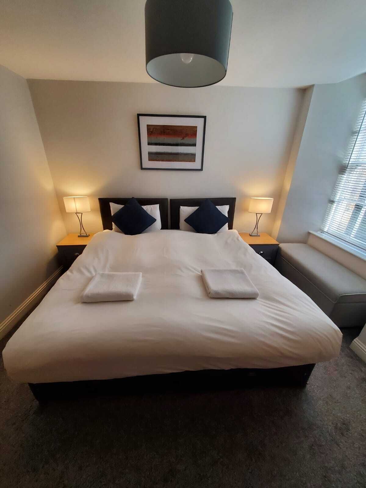 A neatly made bed with white linens and two pillows, a framed artwork on the wall, and two bedside lamps.