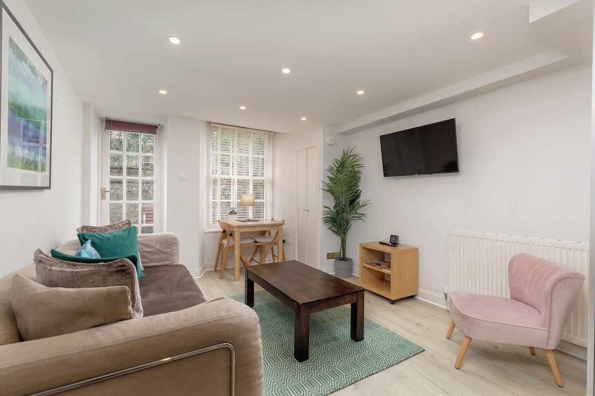 A modern living room with a sofa, a coffee table, a pink armchair, a TV mounted on the wall, and a window with blinds.