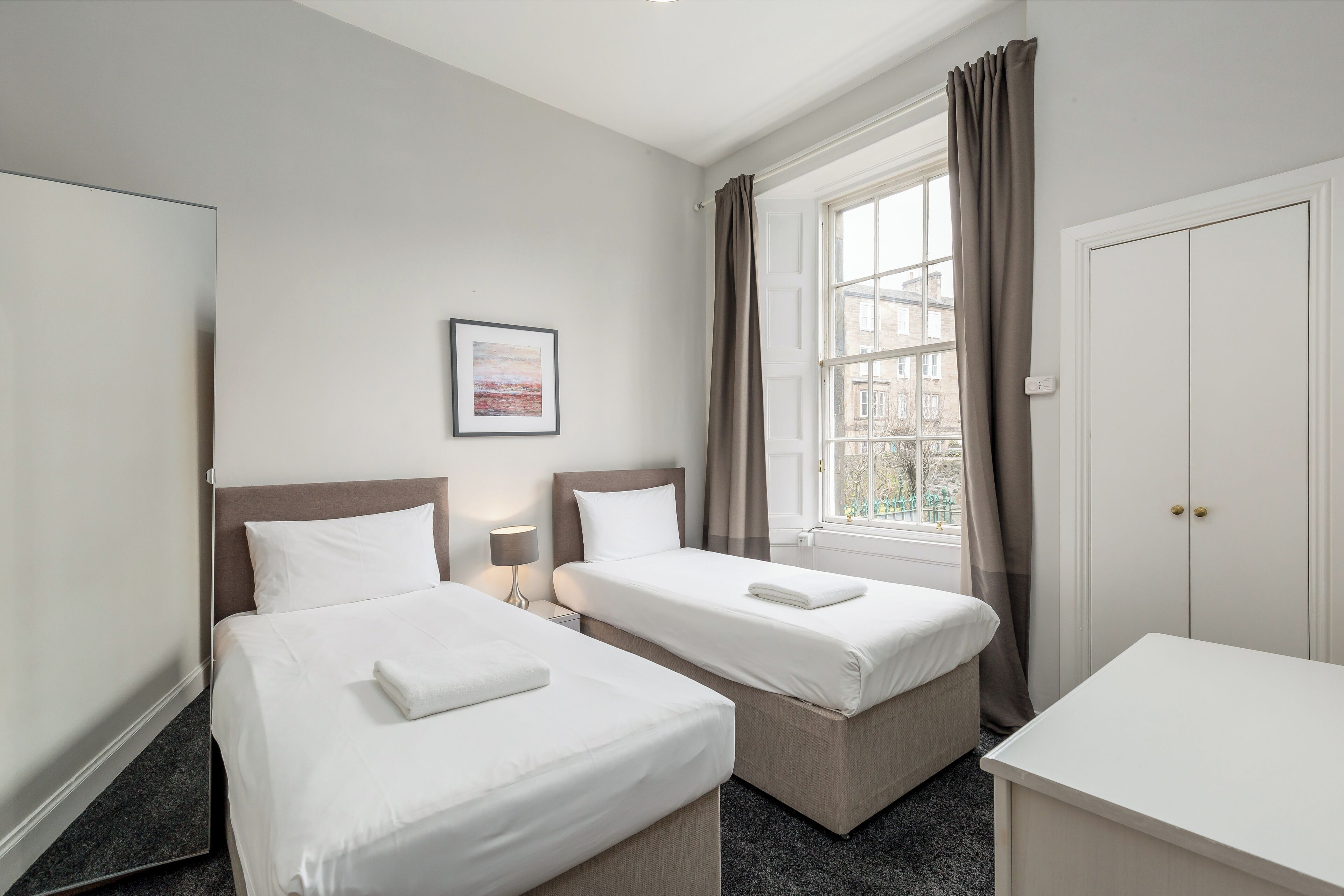 Habitación de hotel con dos camas individuales, una mesita de noche con lámpara, una ventana con cortinas y una puerta.