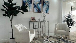 Una sala de estar moderna con dos sillones blancos, una mesa de centro de vidrio, una mesita auxiliar geométrica y arte abstracto en la pared.