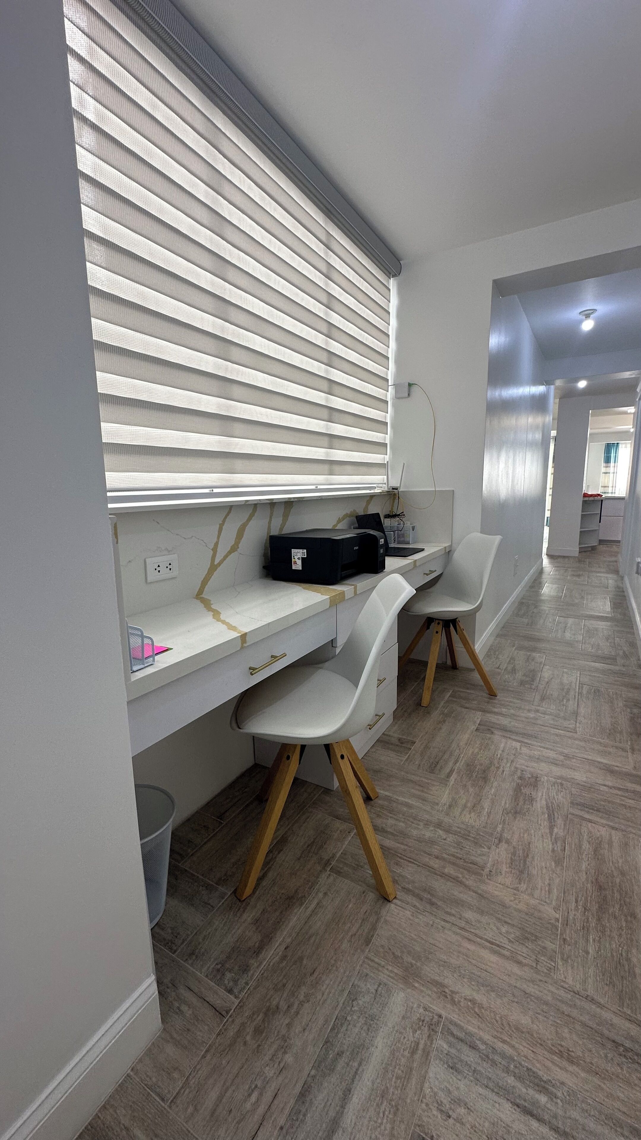A modern office space with a desk, two chairs, a printer, and a window with blinds.