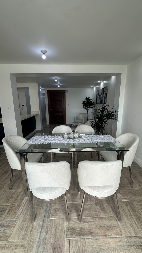 Un comedor con una mesa de vidrio, cuatro sillas blancas tapizadas y una boquilla decorativa.