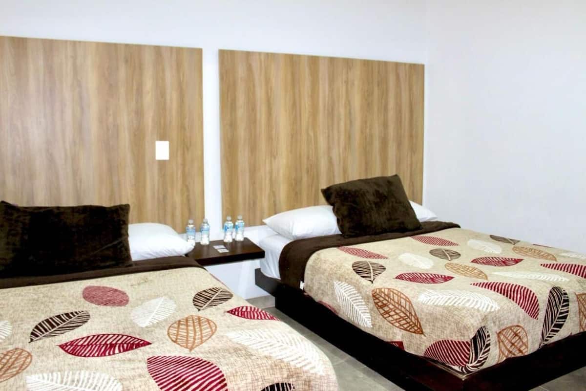 Habitación de hotel con dos camas, cabeceras de madera, una mesita de noche con botellas de agua y una colcha estampada.