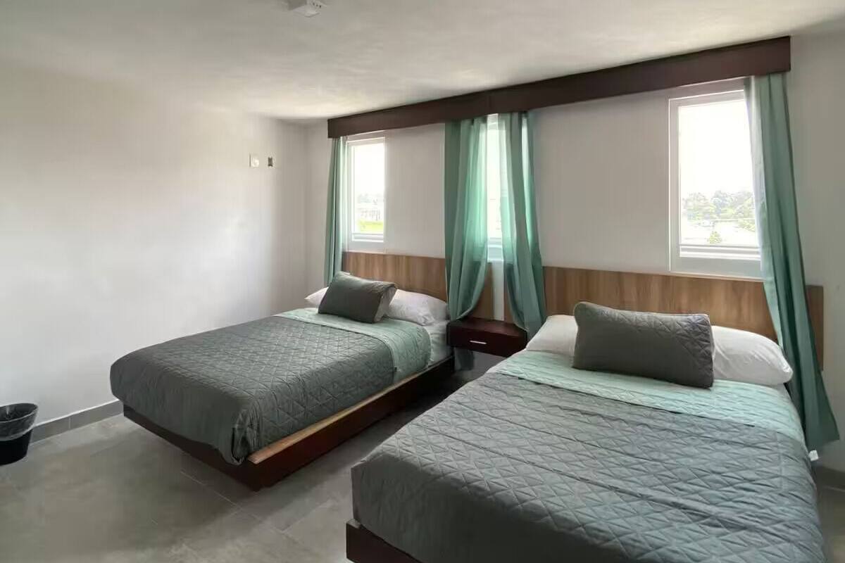 Habitación con dos camas, cada una con cabecera de madera, una mesita de noche pequeña y un velador. Hay una ventana con cortinas, y el piso es de baldosas.