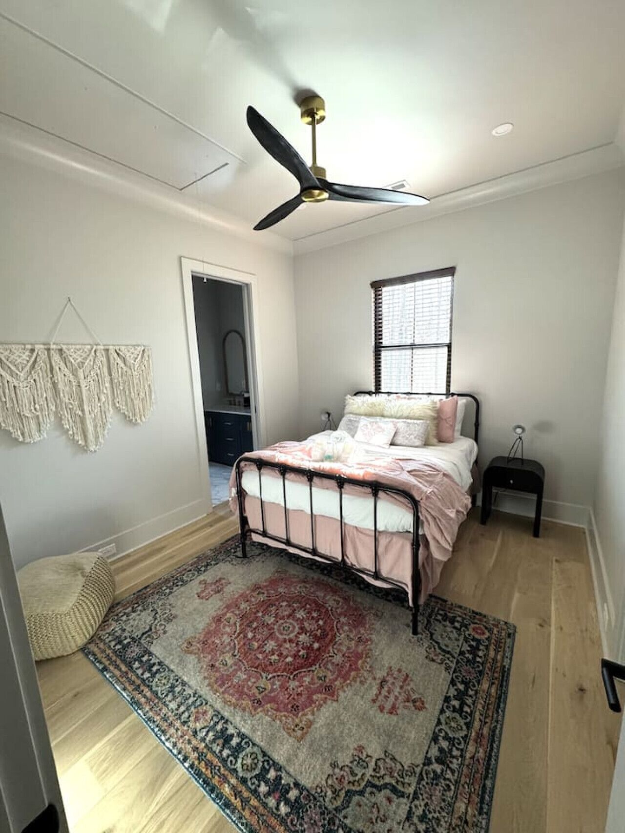 A bedroom with a bed, a ceiling fan, a window with blinds, a rug, and a macrame wall hanging.