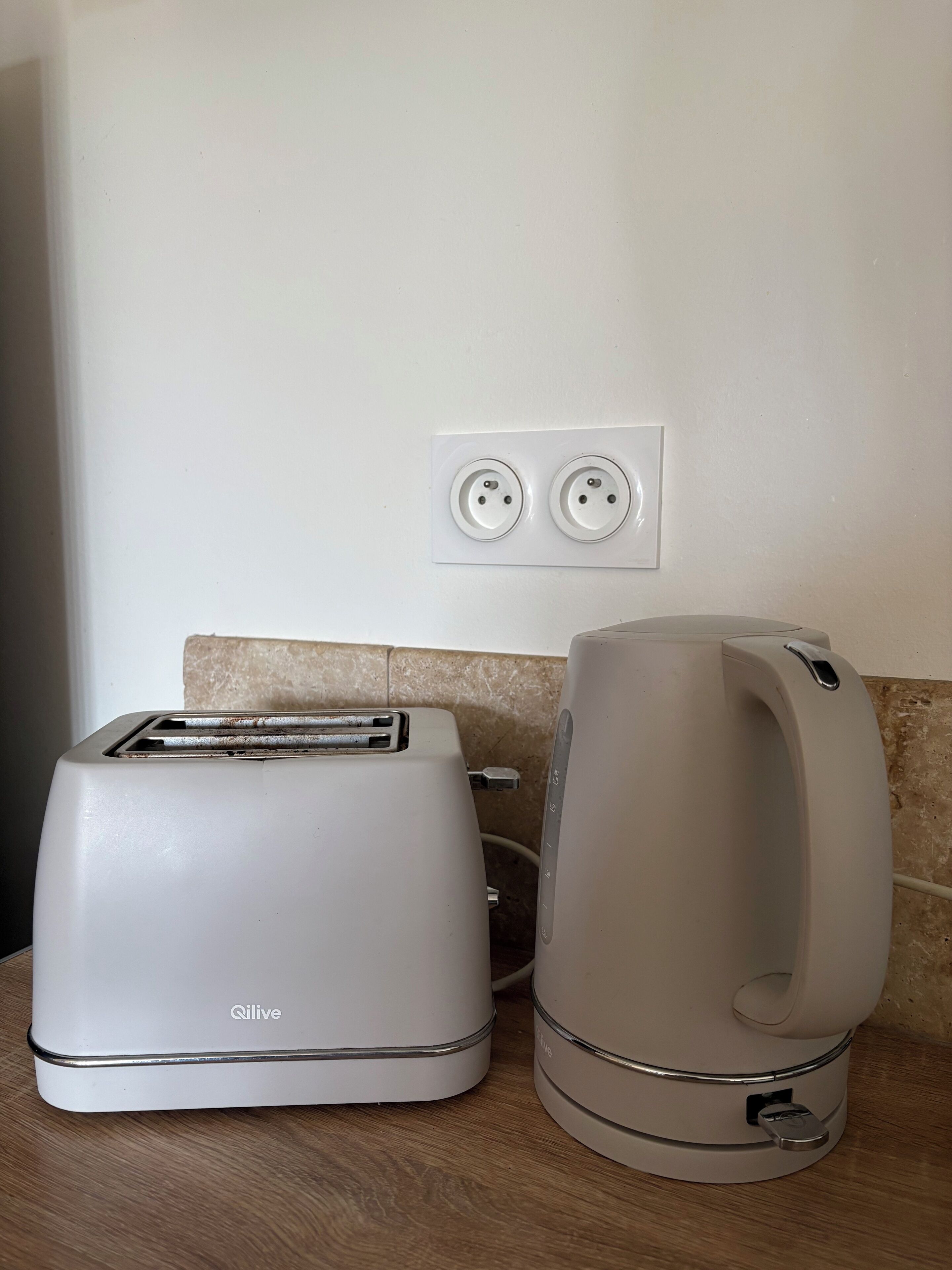 Une cuisine avec un grille-pain blanc et une bouilloire posés sur un plan de travail en bois, une prise électrique au mur et un pare-feu en carreaux.