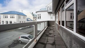 A narrow balcony with a view of a street and parked cars, adjacent to a building with large windows and a metal railing.
