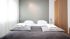 A neatly made double bed with white linens and gray throw pillows, a wooden wardrobe, and a bedside lamp.