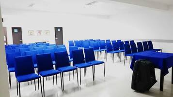 Un salón de conferencias con filas de sillas azules y mesas, un techo blanco y una bolsa negra sobre una mesa.