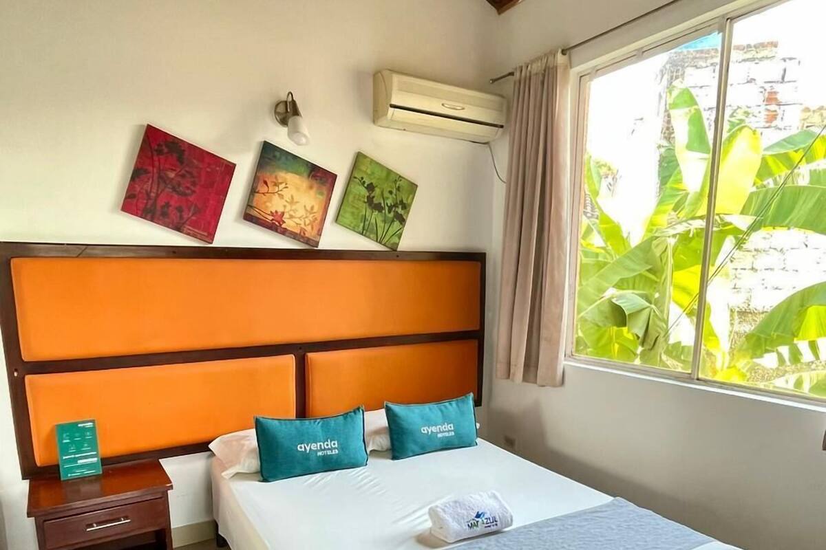 A hotel room with a bed, a nightstand, a window with a view of greenery, and a ceiling with wooden beams.