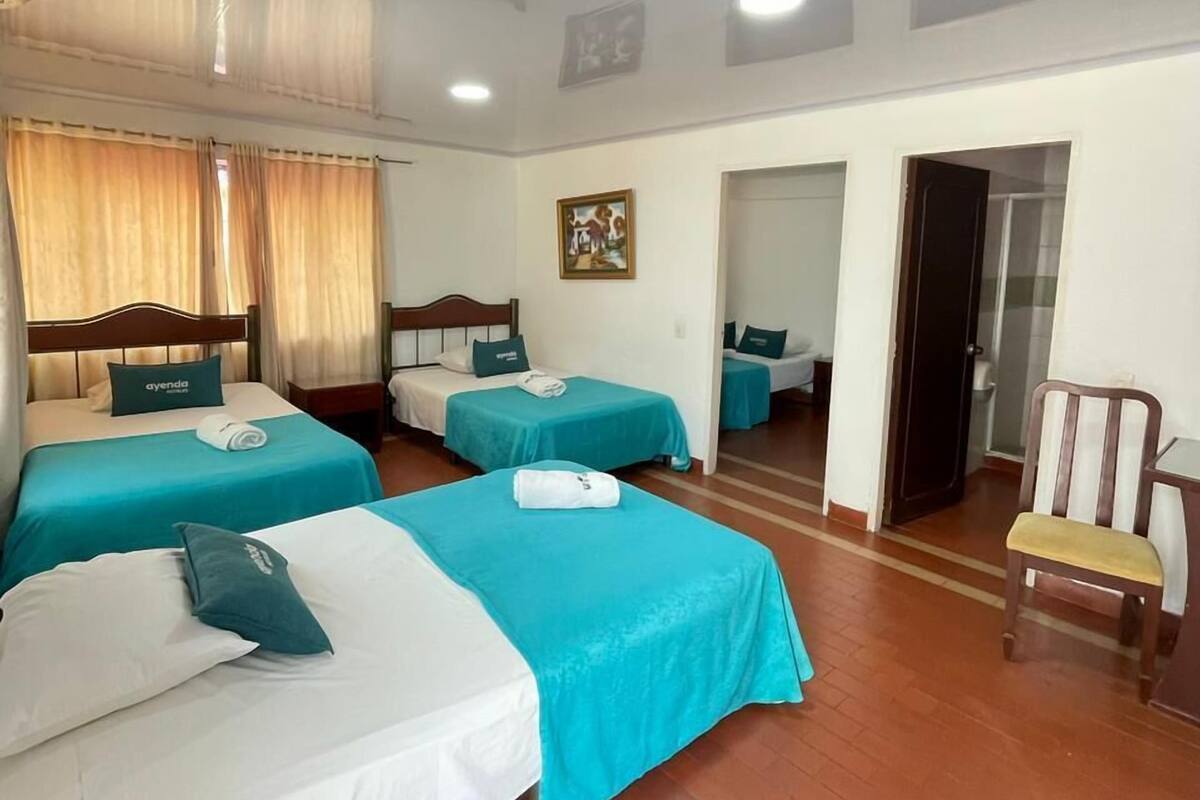 A hotel room with three beds, each with a teal bedspread and white pillows, a wooden chair, and a painting on the wall.