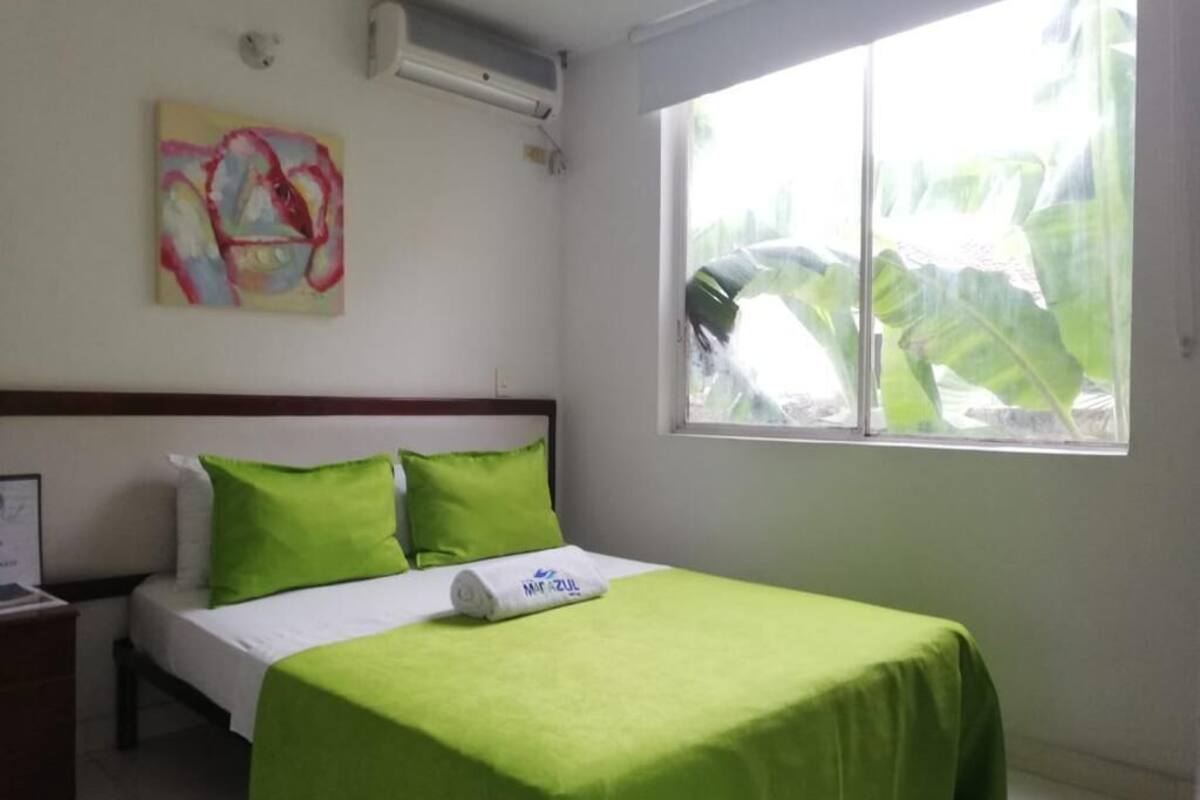 A hotel room with a bed, green bedding, a painting on the wall, and a window with a view of greenery.