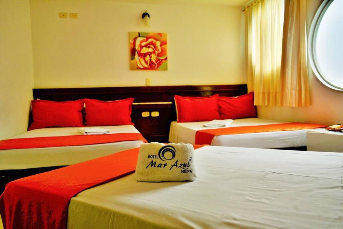 A hotel room with three beds, each with red bedding and pillows. A towel with the hotel name is placed on the bed in the foreground.