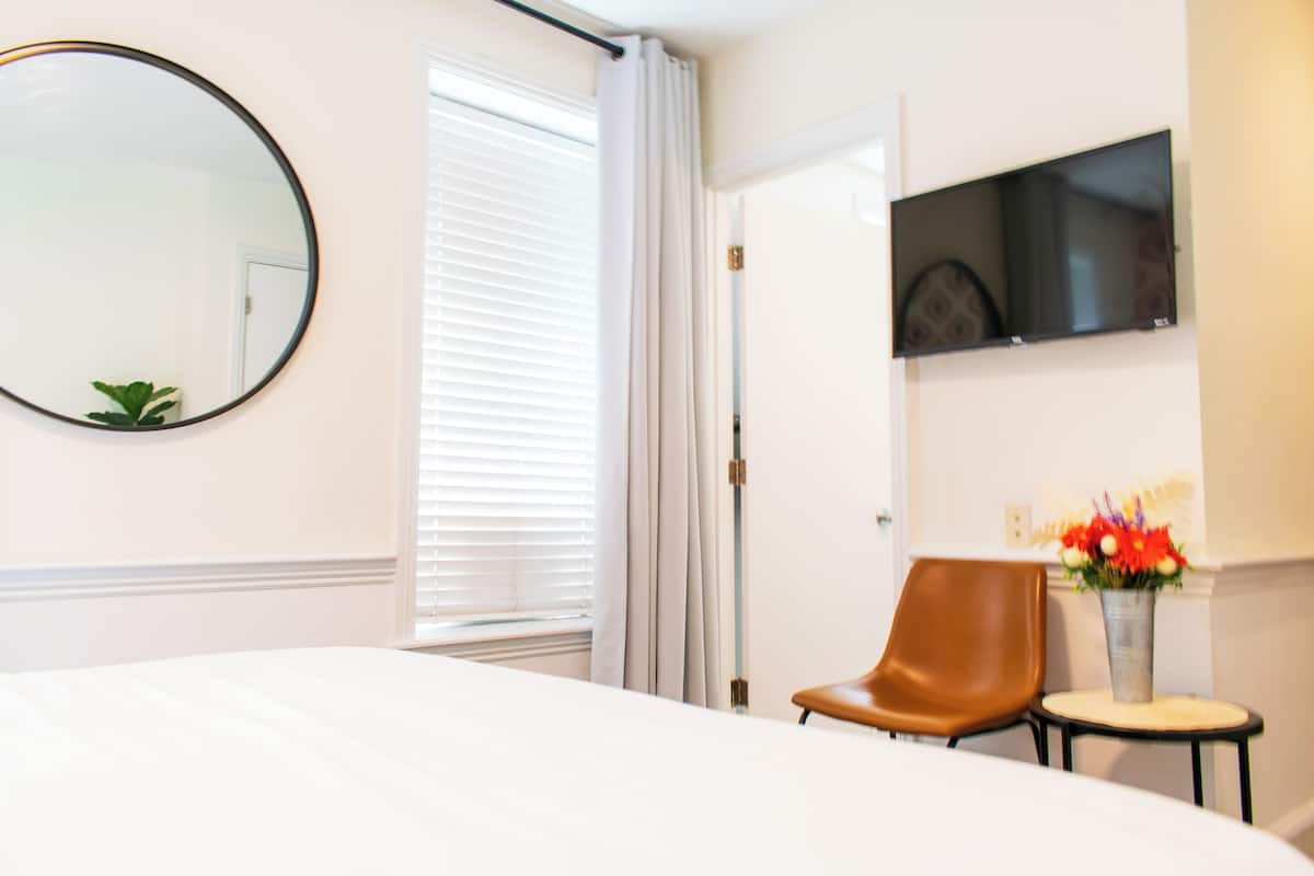 Una camera d'albergo con un letto, una sedia, un tavolino con un vaso di fiori, uno specchio rotondo e una TV appesa al muro.