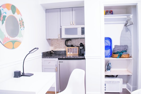 A compact kitchen with a microwave, kettle, and a coffee maker on the countertop. There is a white chair and a small table with a lamp. - Good Hotels Boston (Boston)