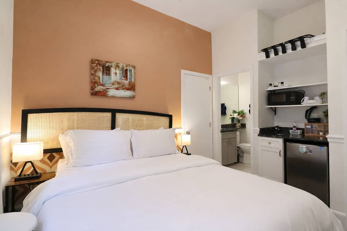 Una camera d'albergo con un letto, lampade sui comodini, un quadro, e un angolo cottura con forno a microonde e frigorifero.