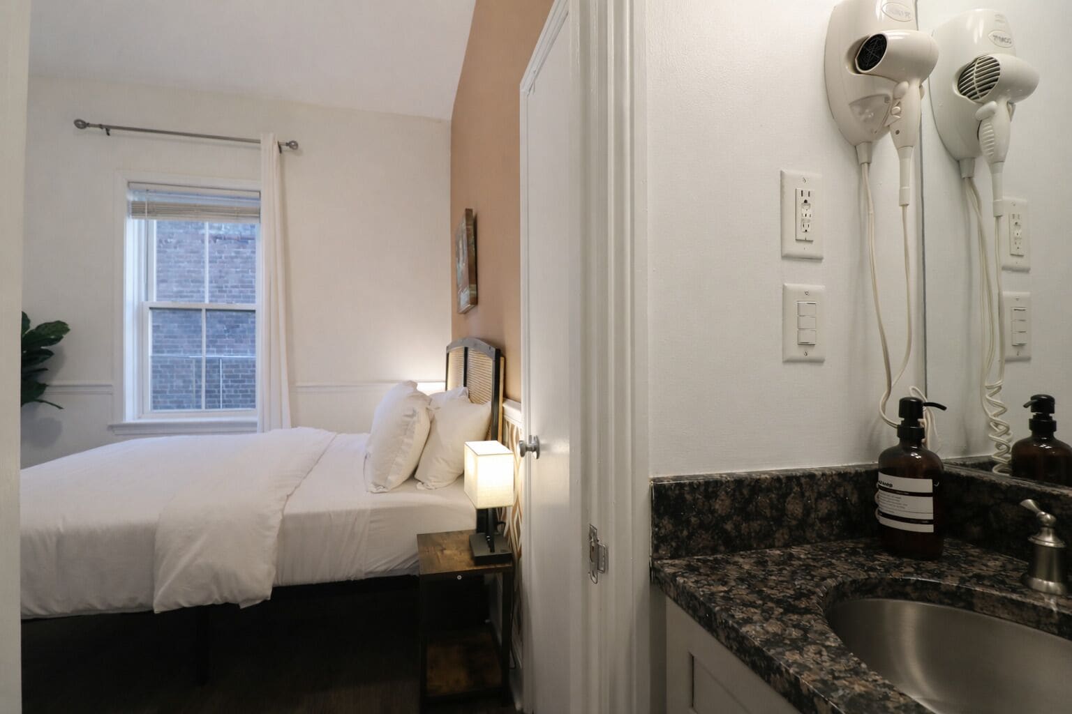 A hotel room with a bed, bedside table, lamp, hair dryers, and a sink.