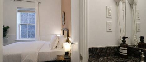 A hotel room with a bed, bedside table, lamp, hair dryers, and a sink.
