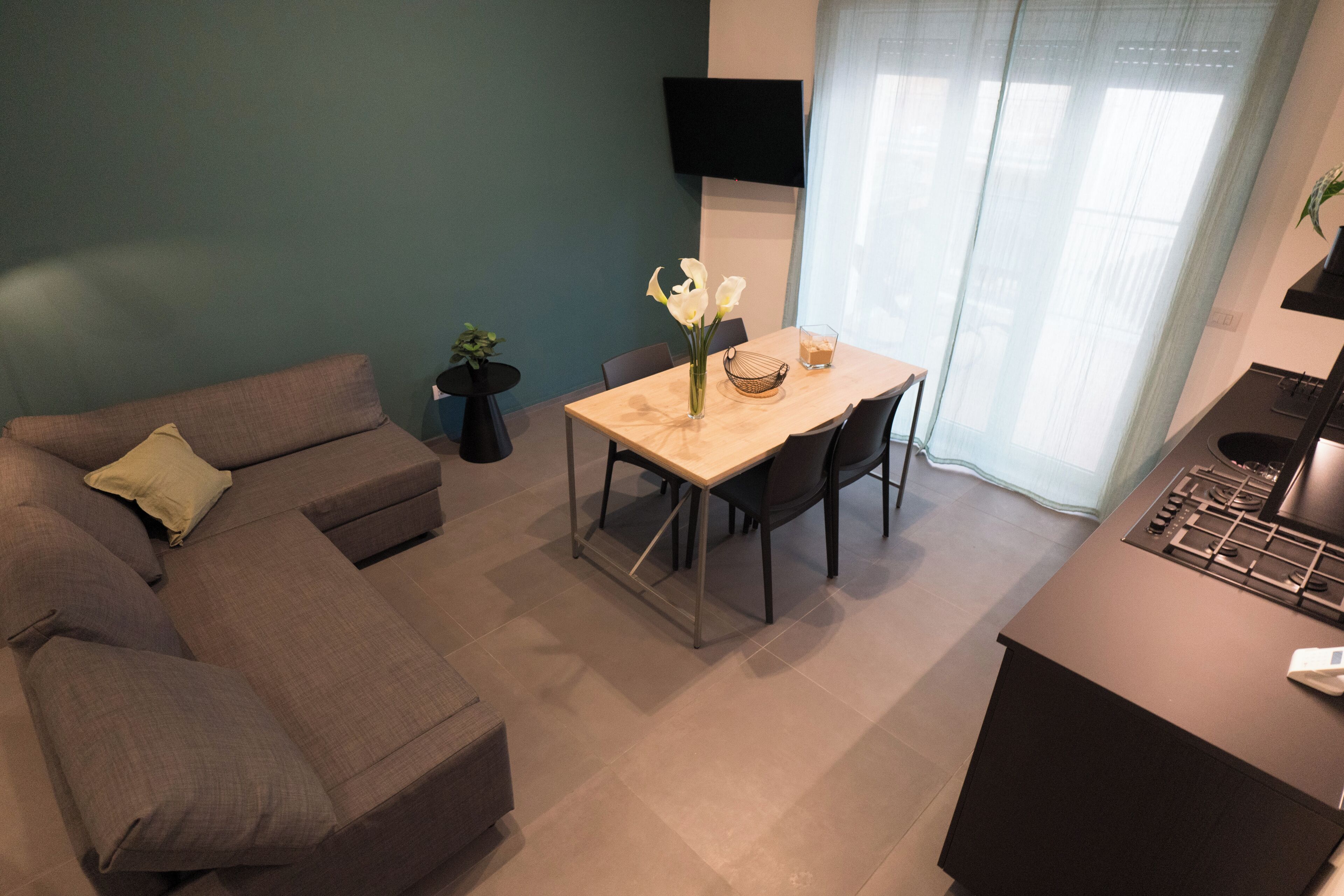 A modern living room with a grey sofa, a dining table with chairs, a small potted plant, and a flat-screen TV mounted on the wall.
