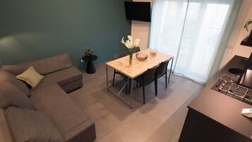 A modern living room with a grey sofa, a dining table with chairs, a small potted plant, and a flat-screen TV mounted on the wall.