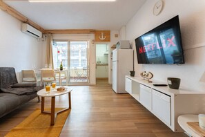 A modern living room with a flat-screen TV, a sofa, a dining area with a table and chairs, and a kitchen area with a refrigerator and microwave.
