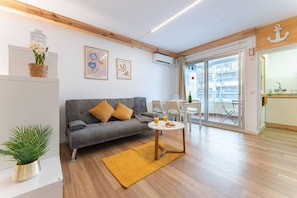 A modern living room with a grey sofa, yellow cushions, a small round table with a bottle and glasses, and a large window offering a view of the city.