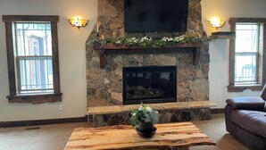 Ein Wohnzimmer mit Kamin aus Stein, einem flachbildfernseher darüber und zwei Fenstern mit dekorativer Beleuchtung.
