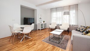 A modern living room with a wooden floor, white furniture, a flat-screen TV, and a large window with curtains. - Comfy Central Apartment (Reykjavík)