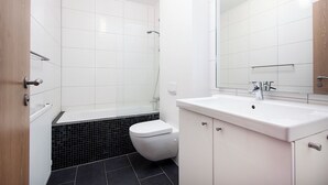 A modern bathroom with white tiles, a black-tiled shower area, a white toilet, and a white sink with a mirror above it. - Comfy Central Apartment (Reykjavík)