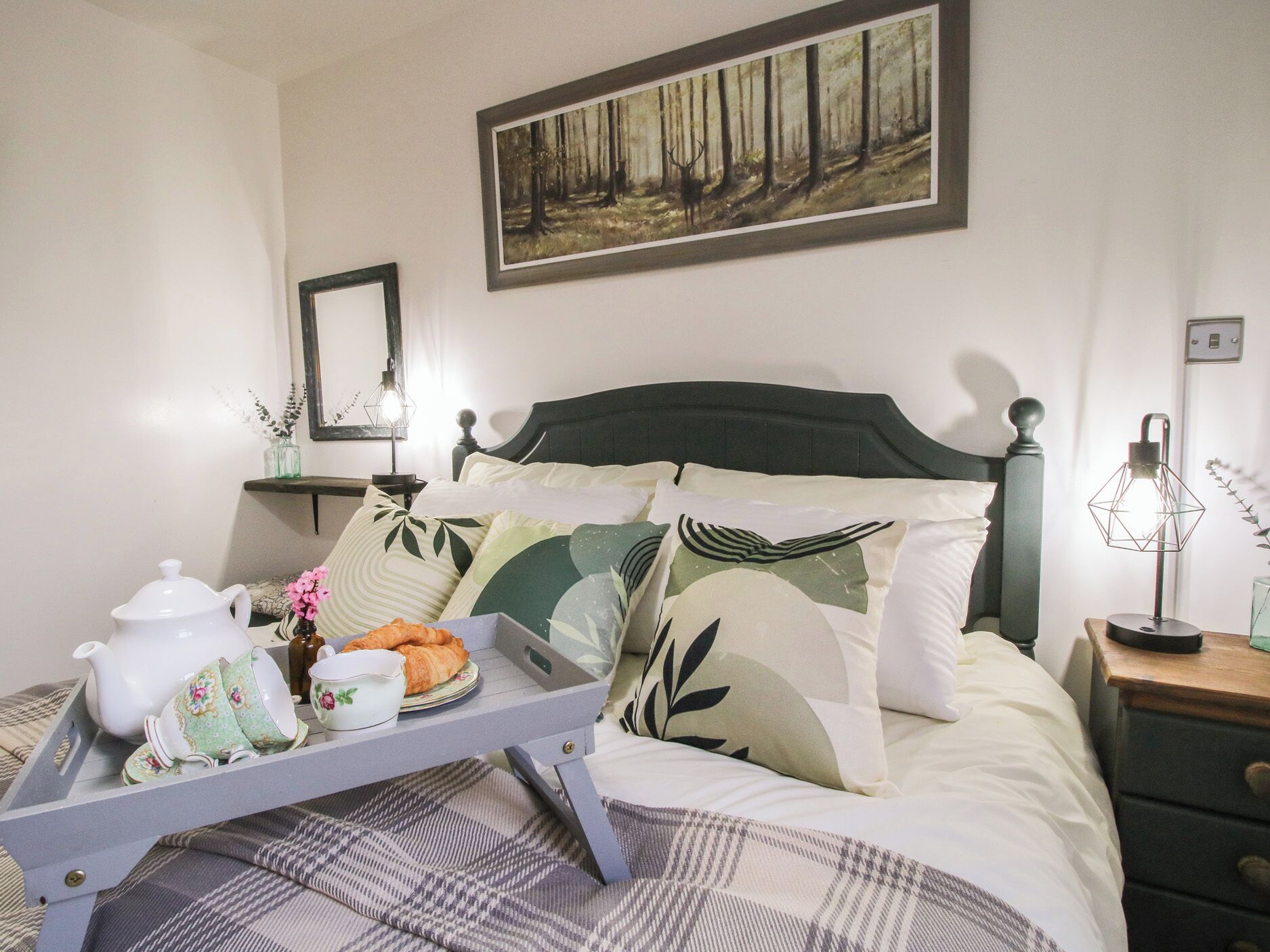 A bedroom with a bed, a bedside table, a tray with breakfast items, a painting on the wall, and a mirror.