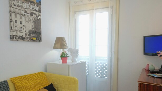 Una sala de estar con un sofá amarillo, una pequeña mesa blanca con una lámpara, una planta en maceta y un televisor sobre un mueble de madera.
