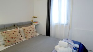 Una cama bien hecha con almohadones estampados, una mesita de noche con lámpara y una ventana con cortinas translúcidas.