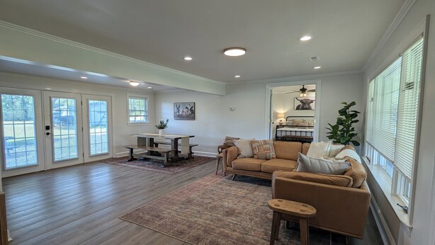 A spacious living room with a wooden floor, a brown sofa, a coffee table, and a dining area with a bench. - Watauga Flats Farm – A Working Farm Stay with Rustic Charm & Modern Comfort! (Johnson City)