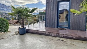 Un edificio moderno con fachada de madera y puerta de vidrio, ubicado sobre una terraza de concreto con vista a montañas al fondo.