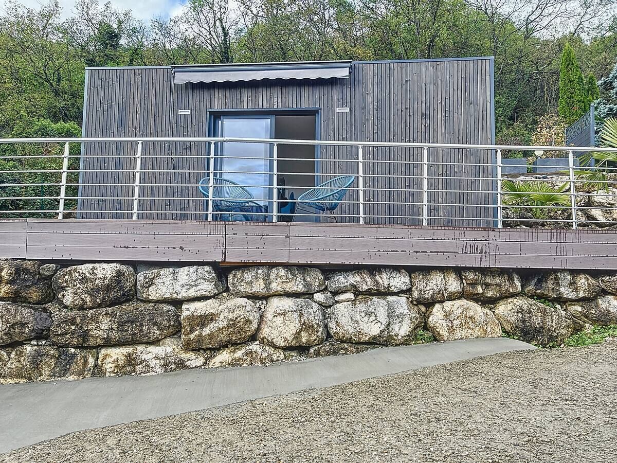 Un bâtiment moderne avec une base en pierre et une rambarde en métal, doté d’une grande porte en verre et de deux chaises bleues à l’extérieur.