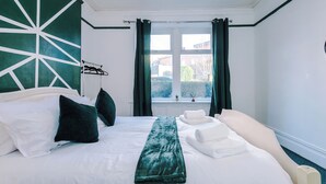 Une chambre d’hôtel avec un grand lit, du linge de lit blanc, un plaid vert et une vue sur l’extérieur par une fenêtre ornée de rideaux.