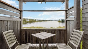 Una terraza de madera con una mesa y dos sillas, con vista a un lago y edificios a lo lejos.